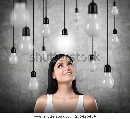 A dreamy brunette looks upward. Lots of light bulbs, one of them is turned on. Concrete background.