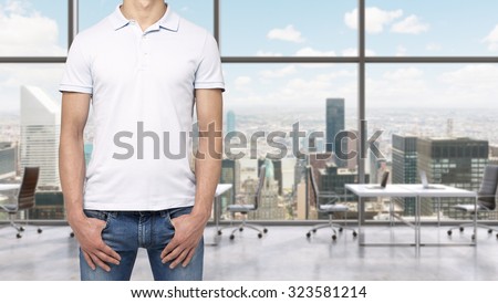 A man in a white polo shirt and denims holds his hands in pockets. A modern panoramic office space in New York City. Manhattan panoramic view.