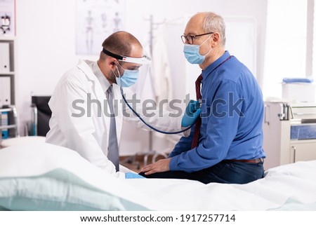 Doctor examining patient lungs using stethoscope wearing face mask as safety precaution in time of covid19. Medical practitioner wearing face mask consulting senior man in examination room during Imagine de stoc © 