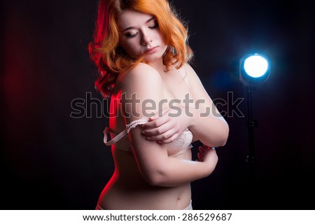 Sexu overweight woman taking off her bra with red and blue light on black background behind her. Studio shooting. Sensuality and sexuality. Beauty and boudoir