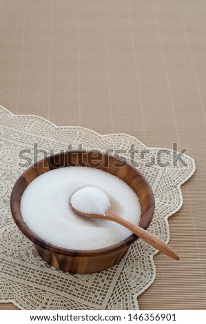 Teaspoon filled with sugar and sugar in wooden bowl.