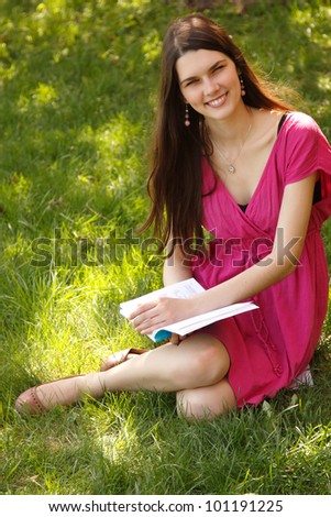 Cheerful attractive student teen girl reading book on green spring grass