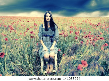 Mysterious portrait of young beautiful woman sitting on stool in a poppy field and looking at camera, summer nature outdoor. Toned.