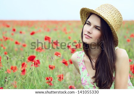 Young beautiful calm girl in straw hat dreams on a poppy field, summer outdoor. Toned, noise added.
