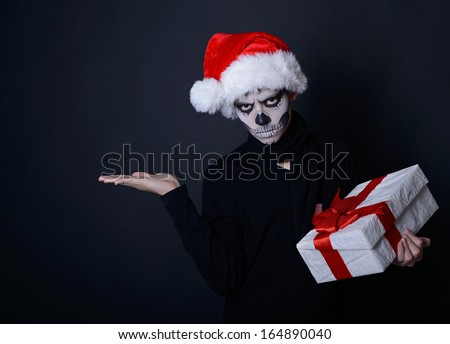 Holiday background of halloween person with terrible skull make-up in santa\'s hat with gift box pointing at empty copyspace over black background