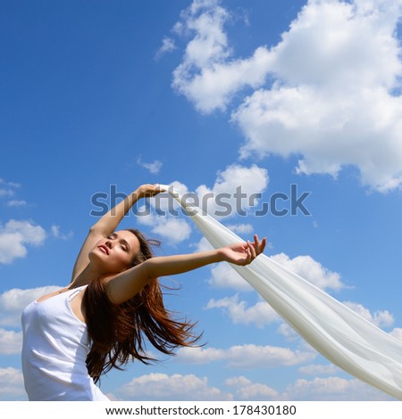 Happy young woman holding white scarf with opened arms expressing freedom, outdoor shot against blue sky.