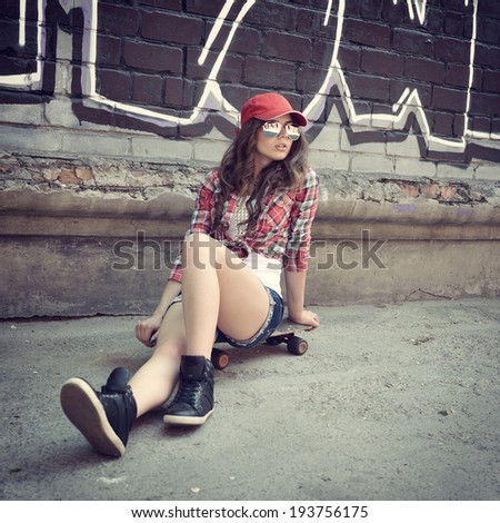Portrait of beautiful teen girl sitting on skateboard over wall with abstract graffiti art. Urban outdoors, teenager\'s lifestyle. Toned.