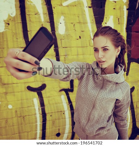 Happy attractive girl with smart phone takes photo of herself against urban grunge graffiti wall, toned.
