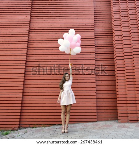 happy young woman standing over red brick wall and holding pink and white balloons