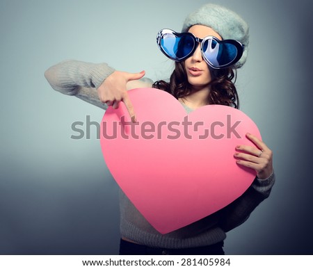 Cute attractive fashion young girl posing with funny big love glasses and pointing at pink heart
