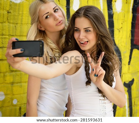 Happy attractive girls with smart phone take selfie against urban grunge graffiti wall.
