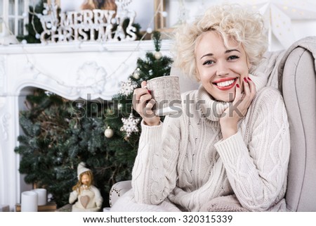 Lovely big smile blonde woman sitting in the cozy comfort of home chair with a blanket wearing a knitted sweater jersey. Series of christmas, x-mas, new year, winter celebration theme photos.