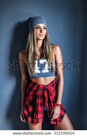 Posing glamor stylish beautiful young woman model in sport shirt in gray hat and Red checkered shirt , red wristlet watch leaned on textured gray wall, looking to the left