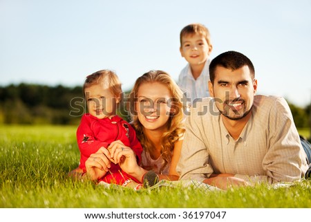 Happy family of 4 people lying on grass under summer sun. Focus is on the man.