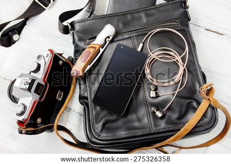 Objects on wooden background: leather bag, camera, smartphone, keys, knife. Outfit of young man.