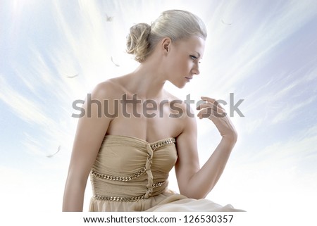 pretty blonde girl with elegant dress sitting on white floor, with a sky as a background , her face is turned is profile at left and her left hand is raised near the shoulder