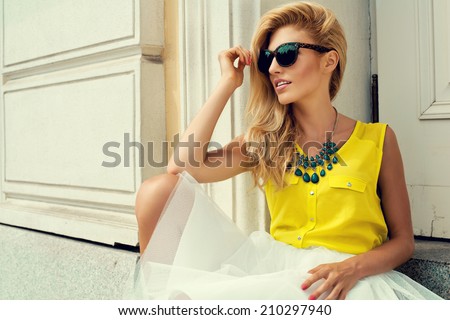 Beautiful blonde young woman holding sunglasses wearing fashionable clothes sitting on stairs