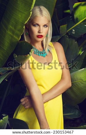 beautiful blonde woman posing in yellow dress and jewelry among palm trees. Fashion photo