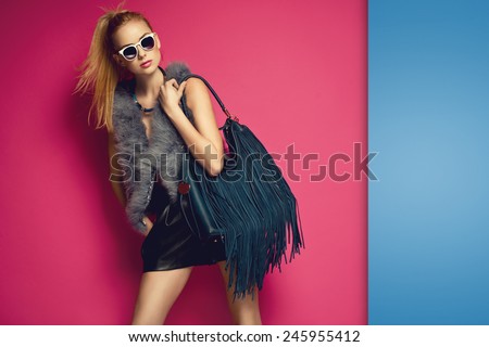 Stylish beautiful blonde woman wearing feather vest holding nice big fringed handbag. Fashion picture