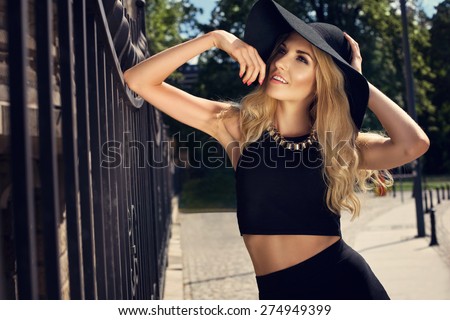Beautiful blonde young woman wearing sunglasses,black top and pencil skirt, hat and handbag, standing on the street. Fashion photo