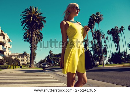 beautiful blonde woman posing with handbag in yellow dress and sunglasses walking across the street. Fashion photo