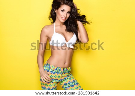Sexy brunette woman wearing white bikini and colorful pants posing on bright yellow background. Perfect body. Skin care and cosmetics