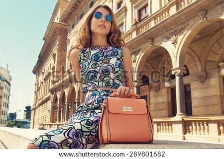 Beautiful blonde young woman wearing fashionable clothes, handbag, sunglasses sitting down in the city. Fashion photo
