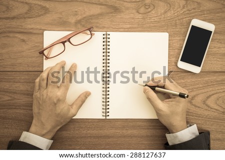 Businessman hand with a pen writing blank notebook on wooden background; retro color effected