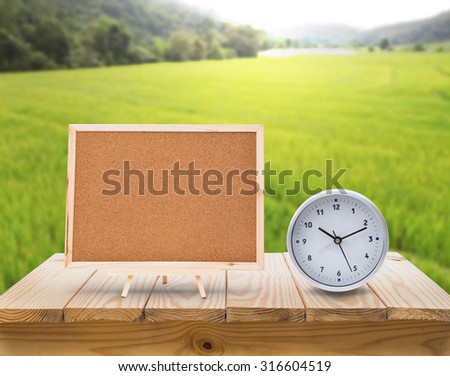 clock and cork board on wood table top with blur green rice fields background
