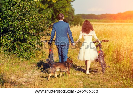 Young happy bride and groom carry bikes in the wheat field back to camera