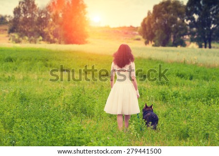 Young bride wearing wedding dress walking with dog on the meadow back to camera.