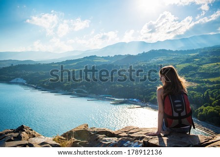 Young tourist smiling woman is sitting on the top of the mounting and with pleasure looking at a beautiful landscape