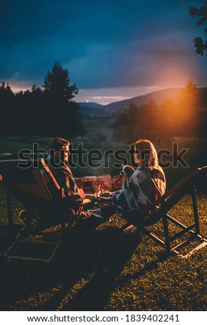 Romantic Couple With Cute Dog Relaxing In Campsite With Fire Pit. Burning Campfire with mountain landscape with night sky over the forest and hills. Getaway in wild nature.