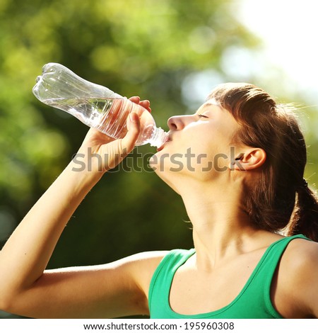 Young sports woman drinking fresh water from the bottle in the sunny park refreshing herself.