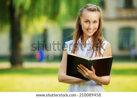 Young happy student woman with the book in her hands is standing in smiling in the university campus. Student is studying in the college yard in fall. Love learning concept.