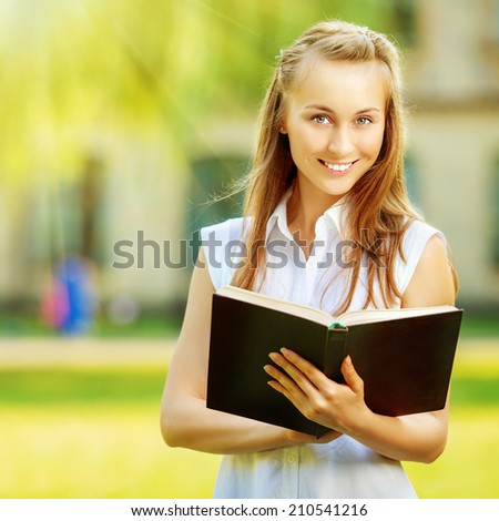 Young happy student woman with the book in her hands is standing in smiling in the sunny university campus. Student is studying in the college yard. Love learning concept.
