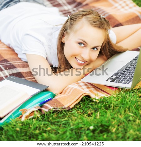 Smiling young student woman is useing a laptop and liying on the grass in the university campusand studying outdoors. Love to study concept.