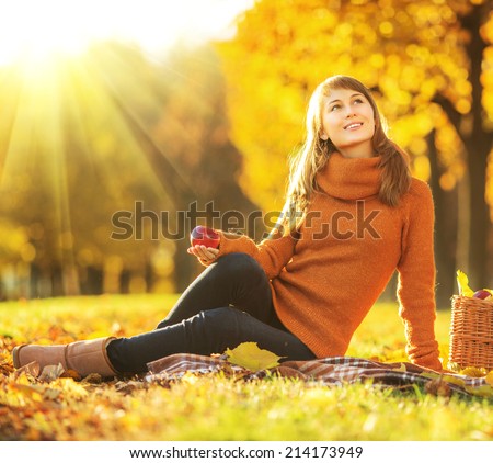 Beautiful young woman is sitting and smiling with rich red apple in her hands and a full basket of tasty rich apples is standing by her on a outdoors autumn sunset background