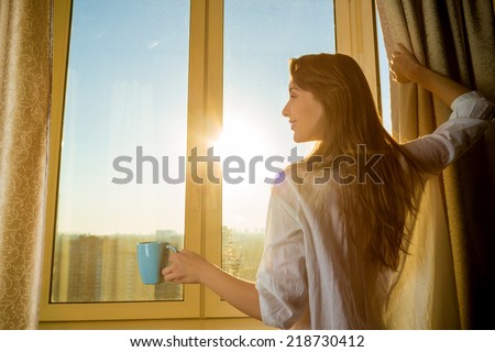 Woman in the morning. Attractive sexy woman with neat body is holding a cup with hot tea or coffee and looking at the sunrise standing near the window in her home and having a perfect cozy morning.