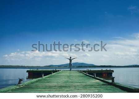 Woman standing on a wooden jetty enjoying the sunshine with arms up. Woman on a pier on the lake greeting the sun with arms wide open.