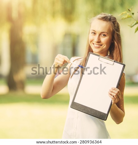 Young woman showing clipboard paperholder on a  green park background. Happy beautiful student woman holding a clipboard with copy space in a sunny spring or summer background in a university campus.