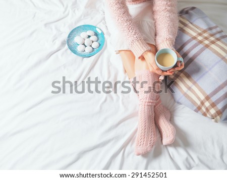 Woman relaxing at cozy home atmosphere on the bed. Young woman with cup of coffee or cocoa in hands and cookies enjoying comfort. Soft light and comfy lifestyle concept.