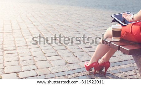 Sexy Business Woman using digital tablet, close. Beautiful woman in red high-heeled shoes and dress enjoying sunny morning in the city, browsing Internet and drinking coffee.