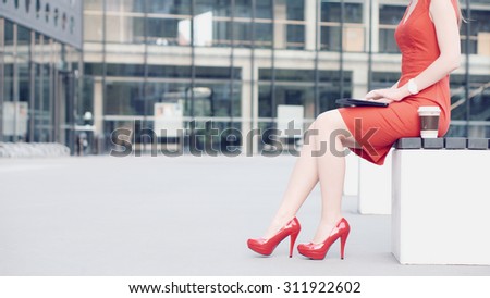 Sexy Business Woman using digital tablet. Beautiful woman in red high-heeled shoes and dress enjoying sunny morning in the city, browsing Internet and drinking coffee.