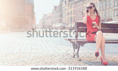Sexy Business Woman talking phone. Beautiful woman in red high-heeled shoes and dress enjoying sunny morning in the city, using smartphone and drinking coffee. Lens Flare.