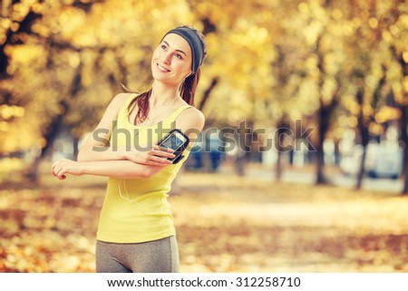 Running woman. Runner is training and listening to music. Female fitness model training outside in the sunny autumn park. Athlete resting in the fall outdoors and using smart phone. Sport lifestyle.