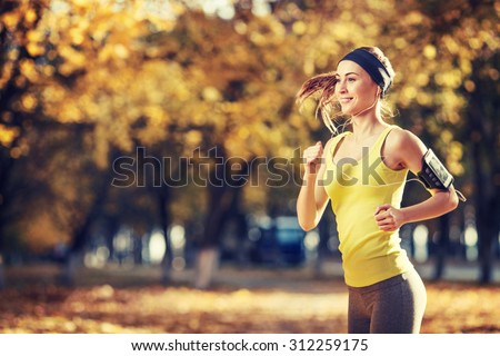 Female fitness model training outside on a warm fall day and listening to music using smart phone. Young woman jogging in autumn park. Sport lifestyle.