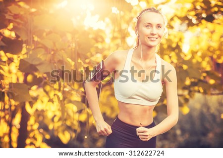 Female fitness model training outside on a warm fall day against sun flare and listening to music using smart phone. Young woman jogging in autumn park. Sport lifestyle.