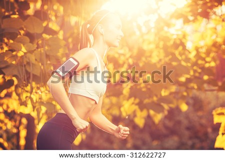 Female fitness model training outside on a warm fall day against sun flare and listening to music using smart phone. Young woman jogging in autumn park. Sport lifestyle.