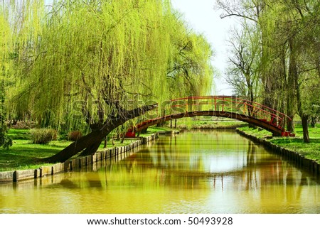 Arched red bridge in a park with willows and water